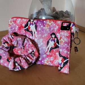 May include: A matching set featuring a pink and purple patterned cosmetic bag, scrunchie, and wristlet. The fabric is decorated with a repeating anime character design and starbursts. A small charm hangs from the wristlet.