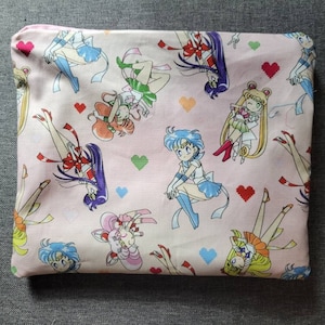 May include: Pink zippered pouch with a repeating pattern of anime characters in various poses. The characters are wearing colorful outfits and have long hair. The background is pink with white hearts.