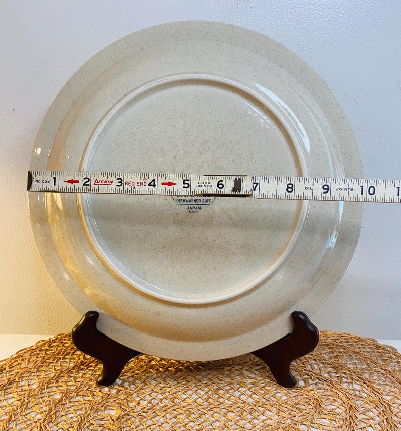 May include: A beige ceramic plate with a speckled design, approximately 25 cm in diameter, is displayed on a dark brown stand. The plate's underside is visible, with text indicating it is dishwasher safe and made in Japan.