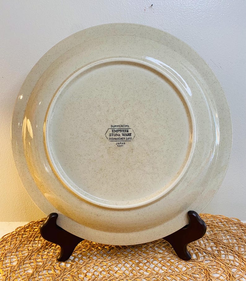 May include: A beige Empress Stone Ware plate with a speckled design. The plate is marked "SUMMERTIME" and "DISHWASHER SAFE" with "JAPAN" printed below. The plate is displayed on a brown stand.