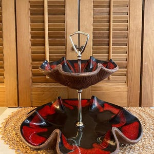 May include: Two-tiered serving dish with a black and red glaze. The top tier is a bowl with a scalloped edge and the bottom tier is a plate with a scalloped edge. The dish has a silver metal stand with a handle.