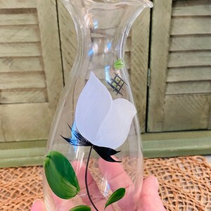 May include: A clear glass vase with a white flower and green leaves painted on the side. The vase has a ruffled top and a rounded body.