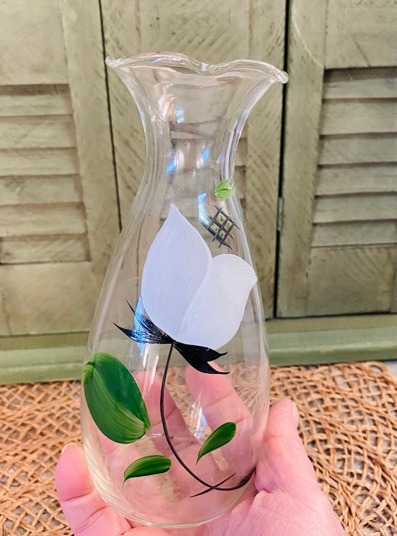 May include: A clear glass vase with a white flower and green leaves painted on the side. The vase has a ruffled top and a rounded body.