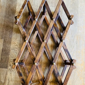 May include: A wooden wine rack with a diamond pattern and brass accents. The rack is folded and has a chain attached to it.