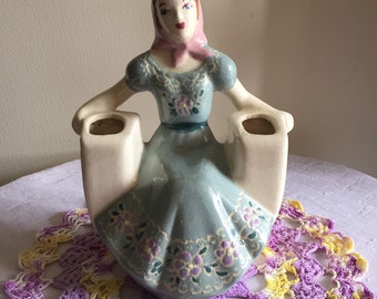 May include: A ceramic figurine of a woman in a blue dress with floral accents, sitting on a bench with two white planters. The woman is wearing a pink scarf and has a painted face.
