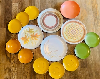 May include: A collection of vintage plates and saucers in various colors and designs. Includes orange, yellow, green, and patterned plates with leaf and geometric designs. The plates are arranged on a wooden surface.