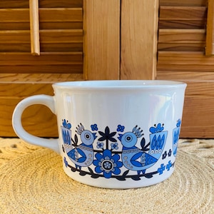 May include: White ceramic mug with a large handle, decorated with a blue bird and floral pattern. The design features stylized birds, flowers, and leaves in shades of blue and black. The mug has a wide rim and a curved handle.