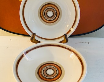 May include: Two white ceramic bowls with brown and orange concentric circle designs. The bowls are stacked on top of each other.
