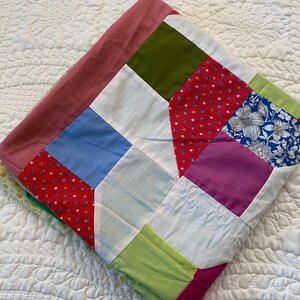 May include: A patchwork quilt with squares of pink, green, red, blue, white, and purple fabric. The quilt is folded in half and is lying on a white surface.