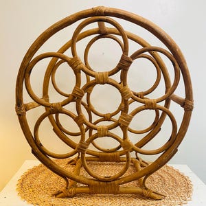 May include: A brown rattan wine rack with a circular design. The rack features multiple circular openings for holding wine bottles. The structure is made of woven rattan and has a natural, rustic appearance. The rack is designed to sit on a flat surface.