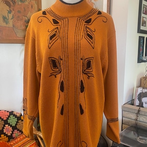 May include: A mustard yellow long-sleeve sweater with a high mock neck collar. The garment features black embroidered geometric and linear patterns on the chest and sleeves. The collar and sleeve cuffs are finished with a thin black trim detail.