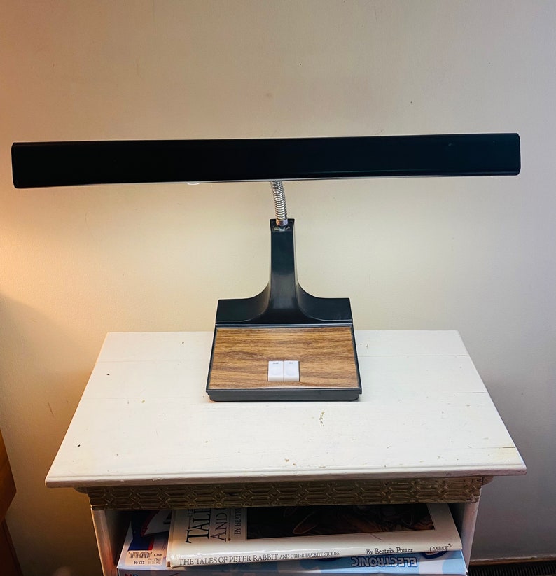 May include: A black desk lamp with a long, rectangular shade and a brown wooden base. The lamp has two white buttons on the base.