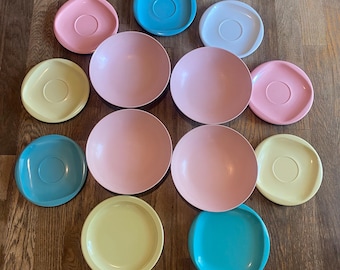 May include: A collection of colorful vintage plates and saucers in shades of pink, blue, and yellow. The plates are round, and the saucers have a central indentation. The arrangement is on a wooden surface, showcasing a retro aesthetic.