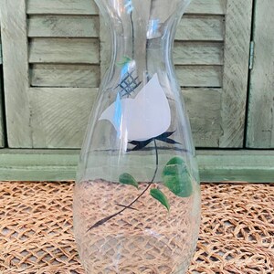 May include: A clear glass vase with a white bird and green leaves painted on the side. The vase has a ruffled top and a rounded bottom.