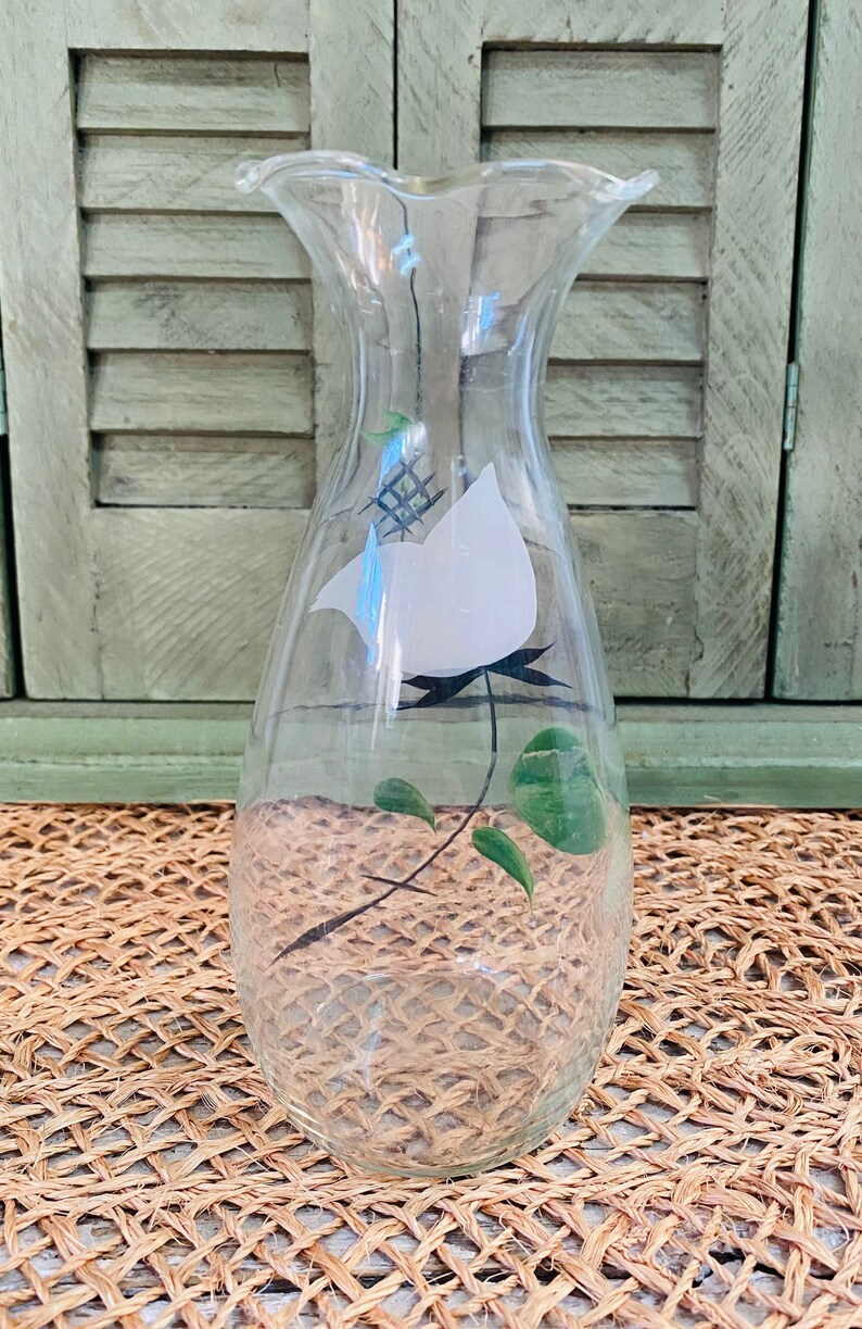 May include: A clear glass vase with a white bird and green leaves painted on the side. The vase has a ruffled top and a rounded bottom.