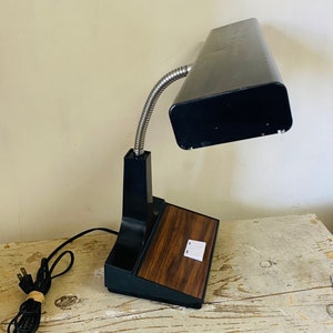 May include: A black desk lamp with a flexible gooseneck and a woodgrain base. The lamp has a rectangular light fixture and two switches on the base.