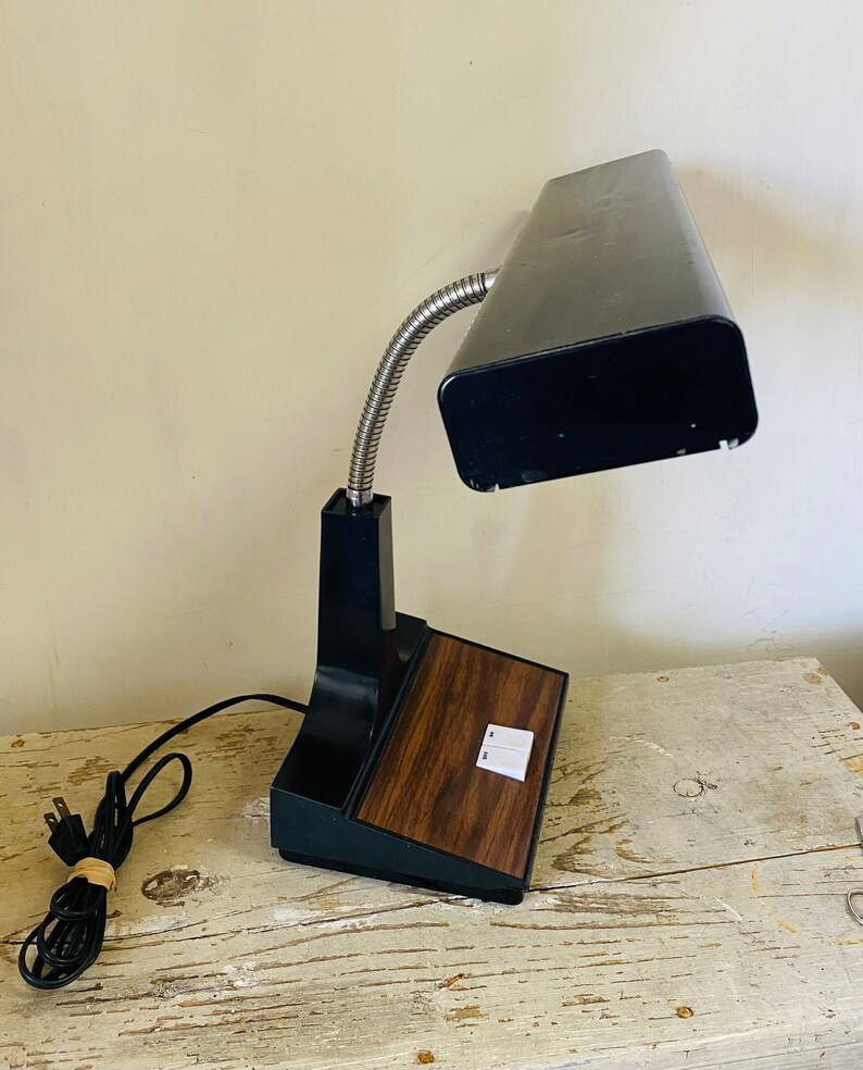 May include: A black desk lamp with a flexible gooseneck and a woodgrain base. The lamp has a rectangular light fixture and two switches on the base.