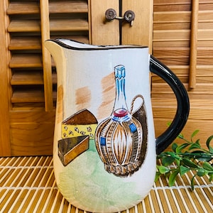 May include: A white ceramic pitcher with a black handle. The pitcher has a painted design of a bottle of wine, two pieces of cheese, and a green background.