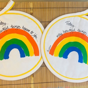 May include: Two white round pot holders with yellow trim. Each pot holder features a rainbow with the text "Today... may beautiful things happen to you..." in black cursive.