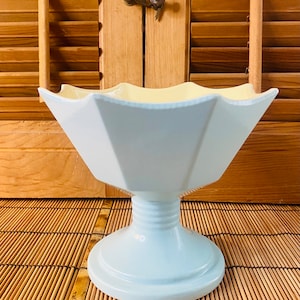May include: A light blue ceramic pedestal bowl with a scalloped rim and a yellow interior. The bowl is on a woven bamboo mat.