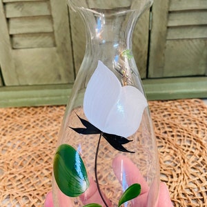 May include: A clear glass bottle with a white flower and green leaves painted on the side. The bottle has a narrow neck and a rounded body.