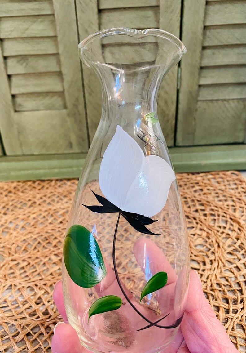 May include: A clear glass bottle with a white flower and green leaves painted on the side. The bottle has a narrow neck and a rounded body.