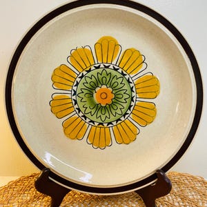 May include: A round ceramic plate with a floral design. The plate has a beige speckled background, a dark brown rim, and a large yellow flower with a green centre. The flower has a small orange centre and black detailing. The plate is on a brown stand.