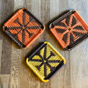 May include: Three woven trivet coasters with geometric patterns. The coasters are made of brown and orange cord, brown and yellow cord, and brown and orange cord. The coasters are square shaped and have a woven design.