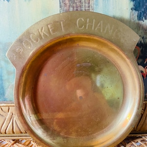 May include: A vintage brass pocket change dish. The dish is round with a curved top that reads "POCKET CHANGE" in capital letters. The brass has a warm, golden hue with some signs of age and use. The dish is designed to hold small items.