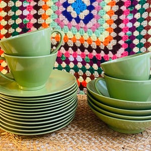 May include: A set of sage green ceramic cups and saucers. The collection includes a tall stack of saucers with two cups placed on top, and a smaller stack of bowls with two additional cups resting on the uppermost bowl, all in a solid color.