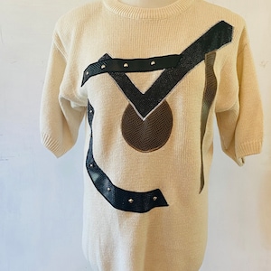 May include: A cream colored short-sleeved sweater with a black and brown geometric design. The design features a circle with a brown center and black leather straps with silver studs.