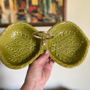May include: A green ceramic double dish with an oak leaf design and a gold metal handle. The dish is perfect for serving snacks or appetizers.