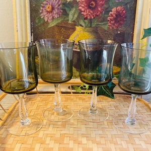 May include: Set of four dark green glass wine glasses with a clear stem and base. The glasses are arranged on a woven wicker surface.