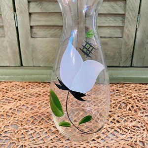 May include: A clear glass vase with a white flower and green leaves painted on the side. The vase has a narrow neck and a wide base.