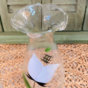 May include: A clear glass vase with a ruffled top and a hand-painted white flower with green leaves and black stem.