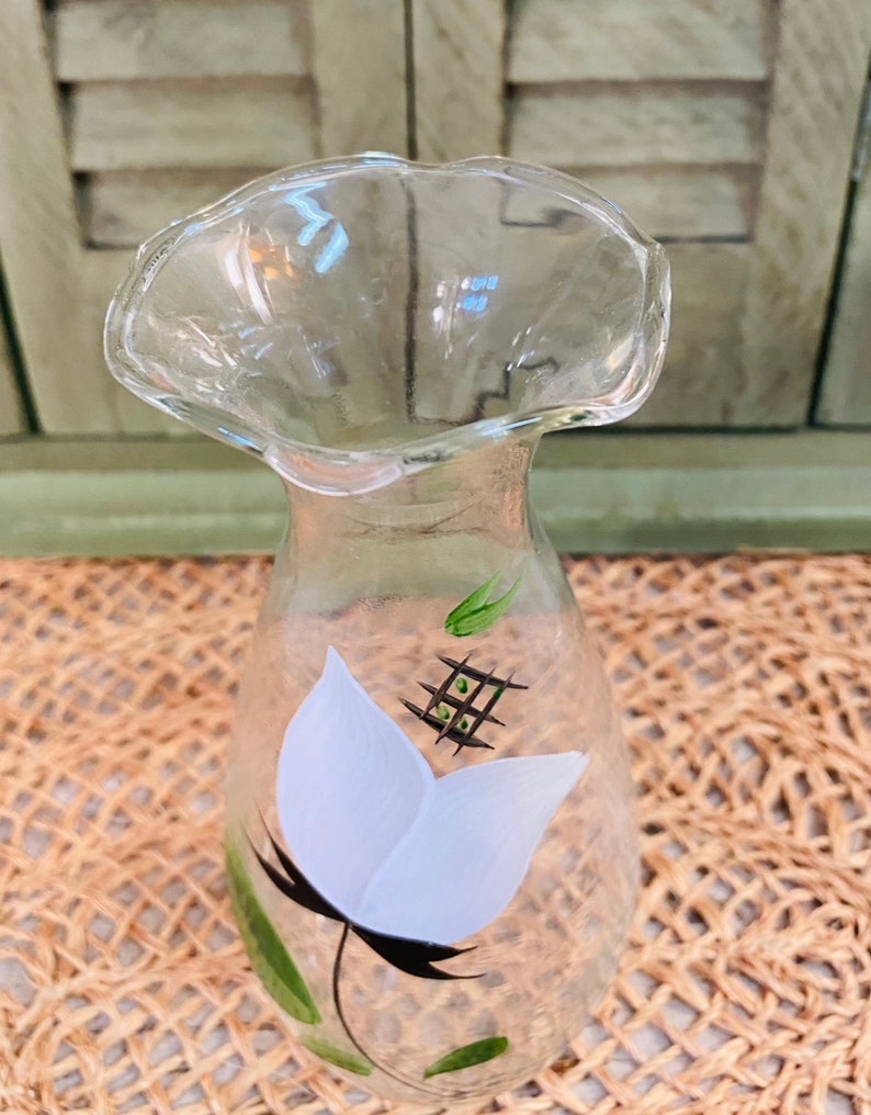 May include: A clear glass vase with a ruffled top and a hand-painted white flower with green leaves and black stem.