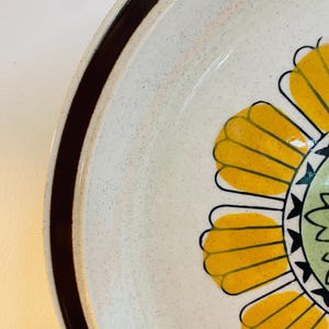 May include: A close-up of a ceramic plate with a floral design. The plate has a cream-coloured speckled background, a brown rim, and a large yellow flower with black outlines. The centre of the flower is green with black accents.