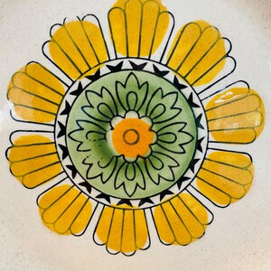 May include: A ceramic plate with a large, stylized floral design. The flower features yellow petals, a green centre, and an orange centre, all outlined in black. The plate has a speckled, off-white background.