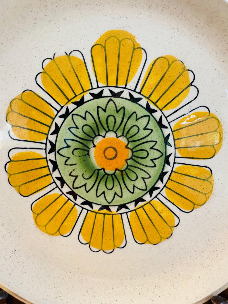 May include: A ceramic plate with a large, stylized floral design. The flower features yellow petals, a green centre, and an orange centre, all outlined in black. The plate has a speckled, off-white background.