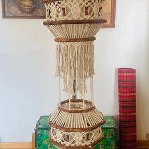 May include: A macrame hanging lamp with two tiers, featuring a wooden frame and white cotton cord. The lamp has a geometric design with tassels and beads.
