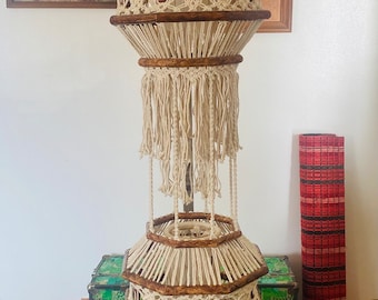 May include: A macrame hanging lamp with two tiers, featuring a wooden frame and white cotton cord. The lamp has a geometric design with tassels and beads.