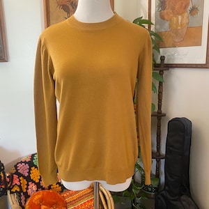 May include: A solid mustard yellow long-sleeve sweater featuring a ribbed crew neckline, ribbed cuffs, and a ribbed hem. The garment is displayed on a mannequin torso, showcasing a simple, plain knit texture throughout the front and sleeves.