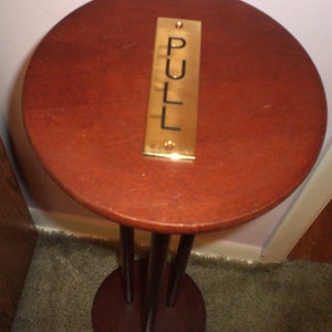 May include: A wooden pedestal with a round top and three legs. The top has a brass plaque with the word "PULL" engraved on it.