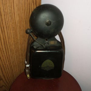 May include: A vintage black electric bell with a large, rounded top and a rectangular base. The bell has a small lever and a cord with a plug. The base features a green and gold emblem. The bell is likely from the early 20th century.