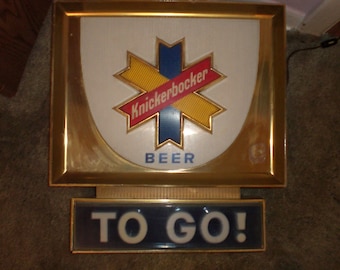 Puede incluir: Un letrero iluminado vintage para la cerveza Knickerbocker. El letrero tiene un marco dorado y presenta una estrella azul, amarilla y roja con las palabras "Knickerbocker Beer" sobre un fondo blanco. El letrero también tiene un letrero más pequeño debajo que dice "TO GO!" en letras blancas sobre un fondo azul.