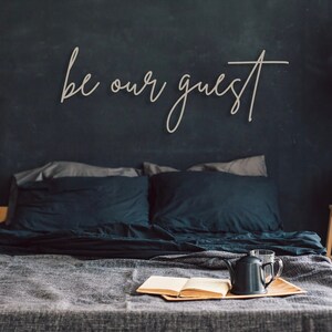 May include: A wall-mounted wooden sign featuring the cursive text be our guest in a light tan finish. The sign is displayed above a bed with dark bedding, a wooden tray holding an open book and a black teapot, a striped laundry basket, and a side table.