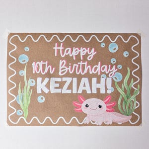 May include: A brown paper birthday card with the text "Happy 10th Birthday KEZIAH!" in pink and white. The card features a cartoon axolotl, bubbles, and seaweed illustrations. The card is decorated with a white wavy border.