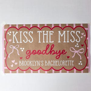 May include: A rectangular sign with the text "KISS THE MISS goodbye" in white and red lettering. The sign is brown with a pink scalloped border and pink accents, including hearts and bows. The text "BROOKLYN'S BACHELORETTE" is also included.