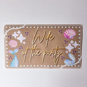 May include: A rectangular sign with the words "Wife of the party" in gold script. The sign is brown with white pearl and dot borders, and features illustrations of seashells, starfish, and a mermaid tail.