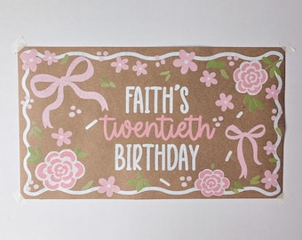 May include: A rectangular brown paper sign featuring the white and pink text "FAITH'S twentieth BIRTHDAY". The design includes a white scalloped border, light pink bows, and pink floral illustrations with small green leaves scattered throughout.